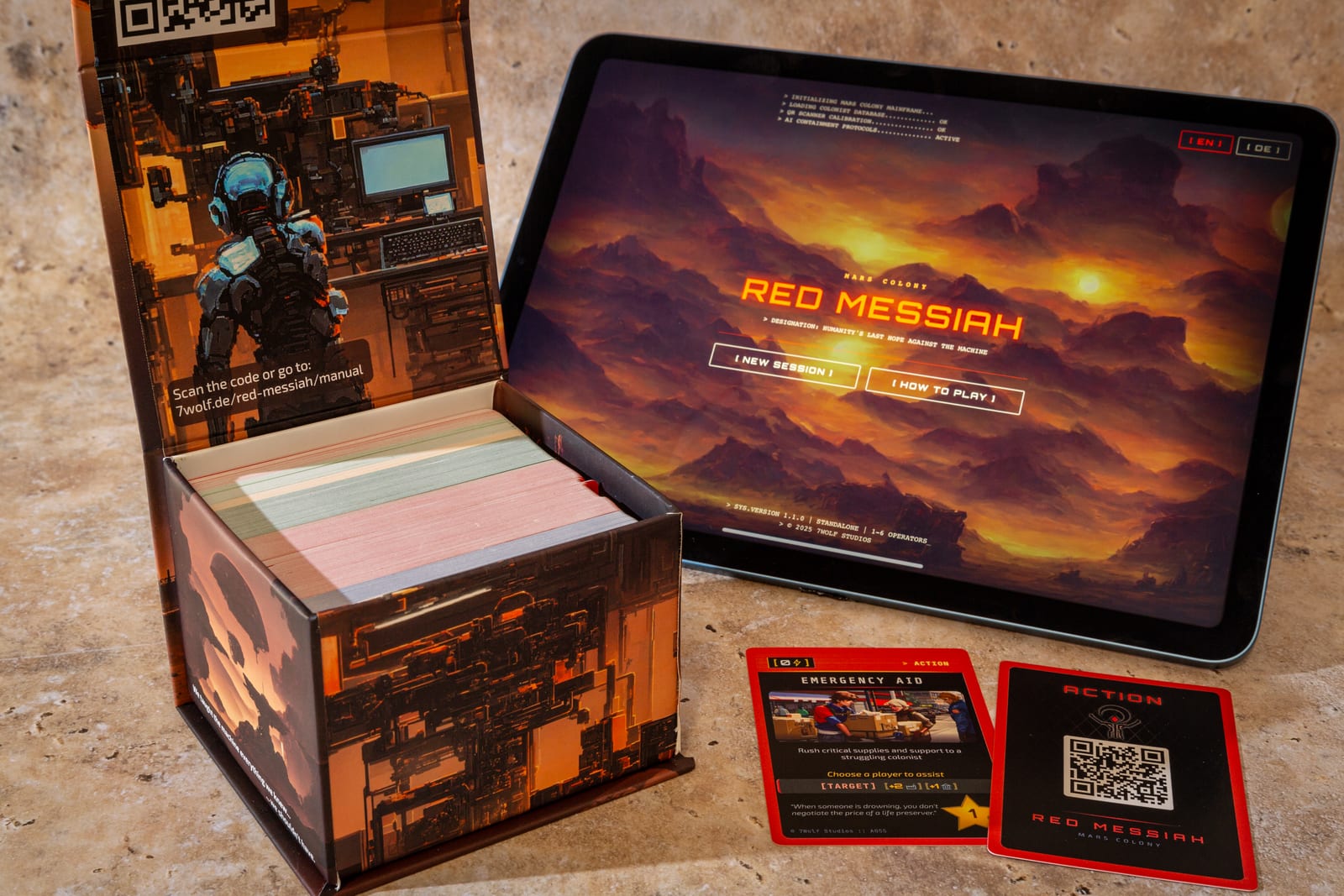 Red Messiah on a table: the open card box with four colour-coded decks, a tablet showing the Mars Colony start screen, and an Emergency Aid action card next to its QR-coded back