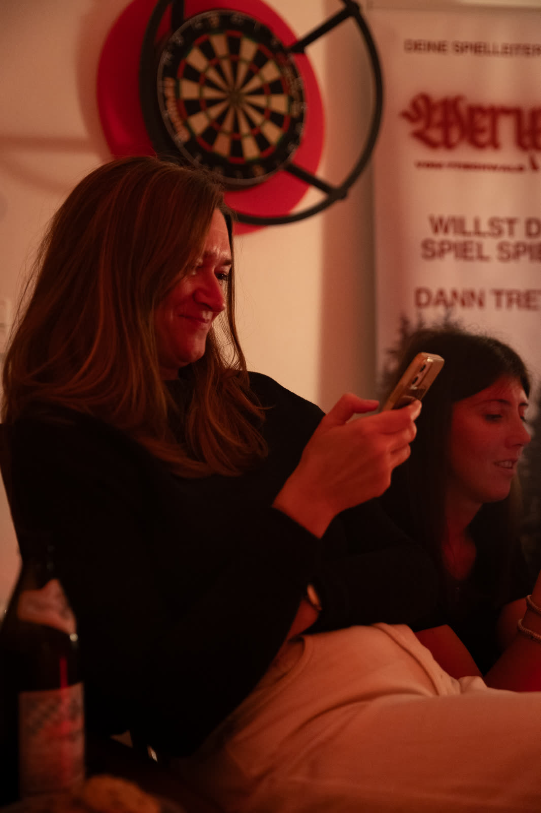 Zwei Spielerinnen am Tisch schauen lächelnd auf ihr Handy, dahinter ein Werwolf-Banner